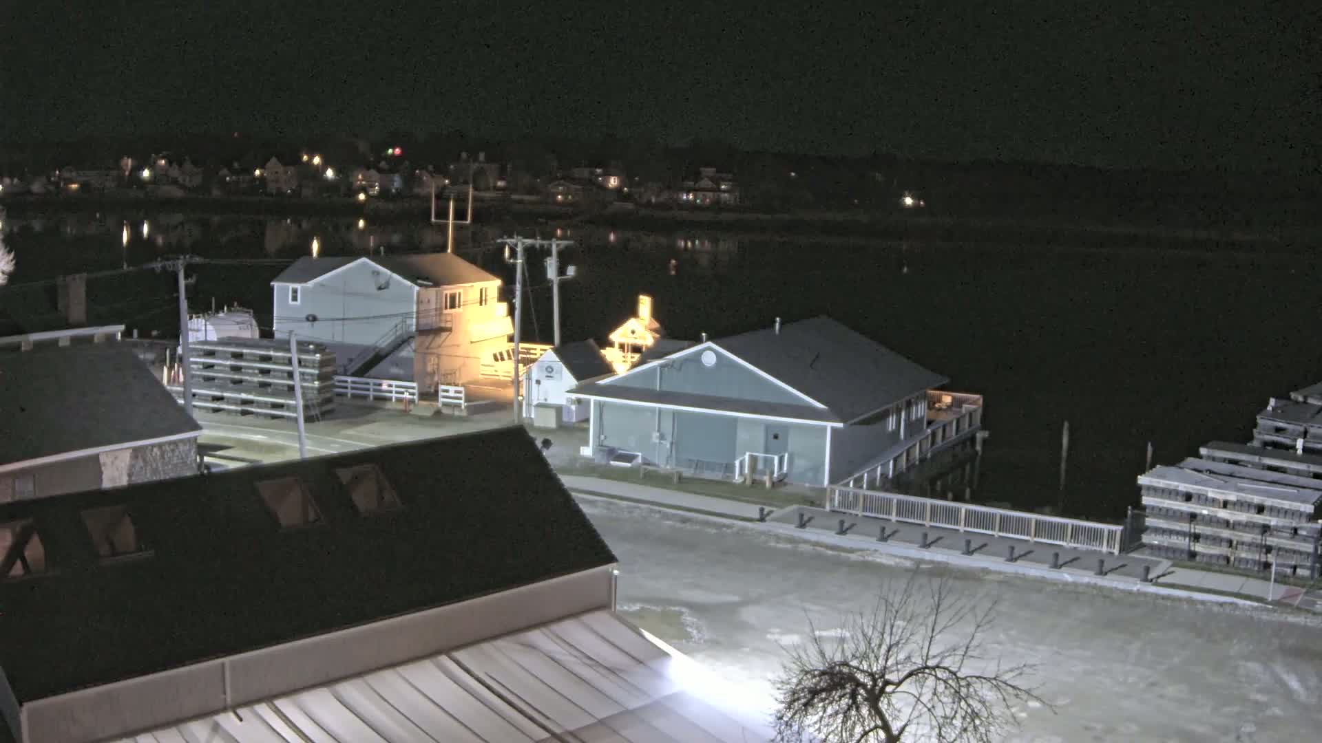A nighttime view of a waterfront town with several buildings illuminated near a calm body of water, stacks of what appear to be lobster traps, and a few bare trees.