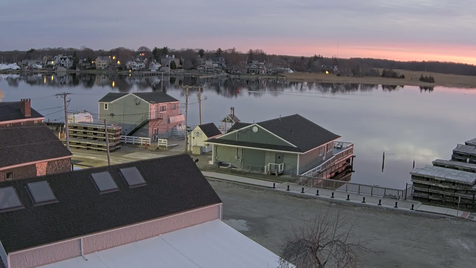 A waterfront town at dusk, with calm water reflecting the pale pink and purple sunset sky, shows several buildings near a dock with stacked wooden structures.