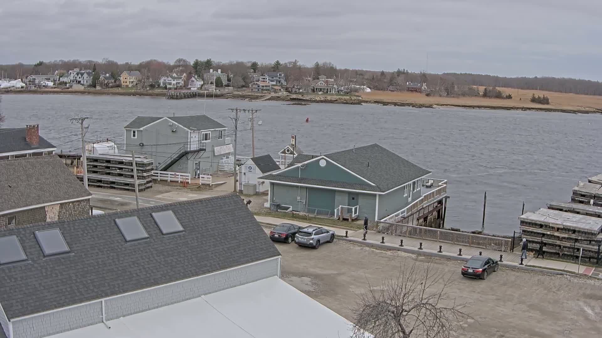 A waterfront scene on an overcast day shows several buildings, a parking lot with cars, and a body of water with a town visible in the distance.