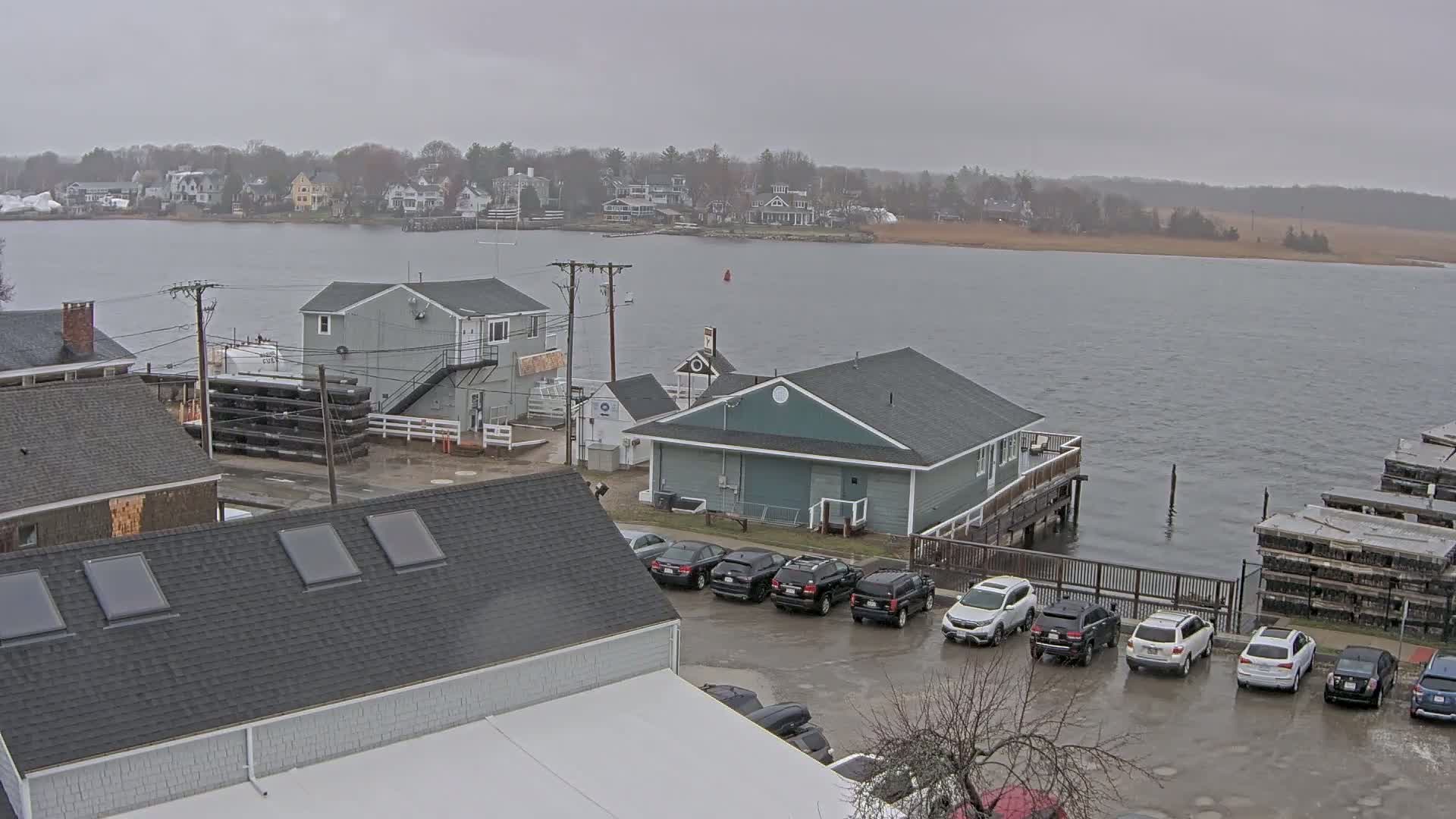 A waterfront town on an overcast day shows several buildings, cars parked in a lot near a body of water, and a distant shoreline.