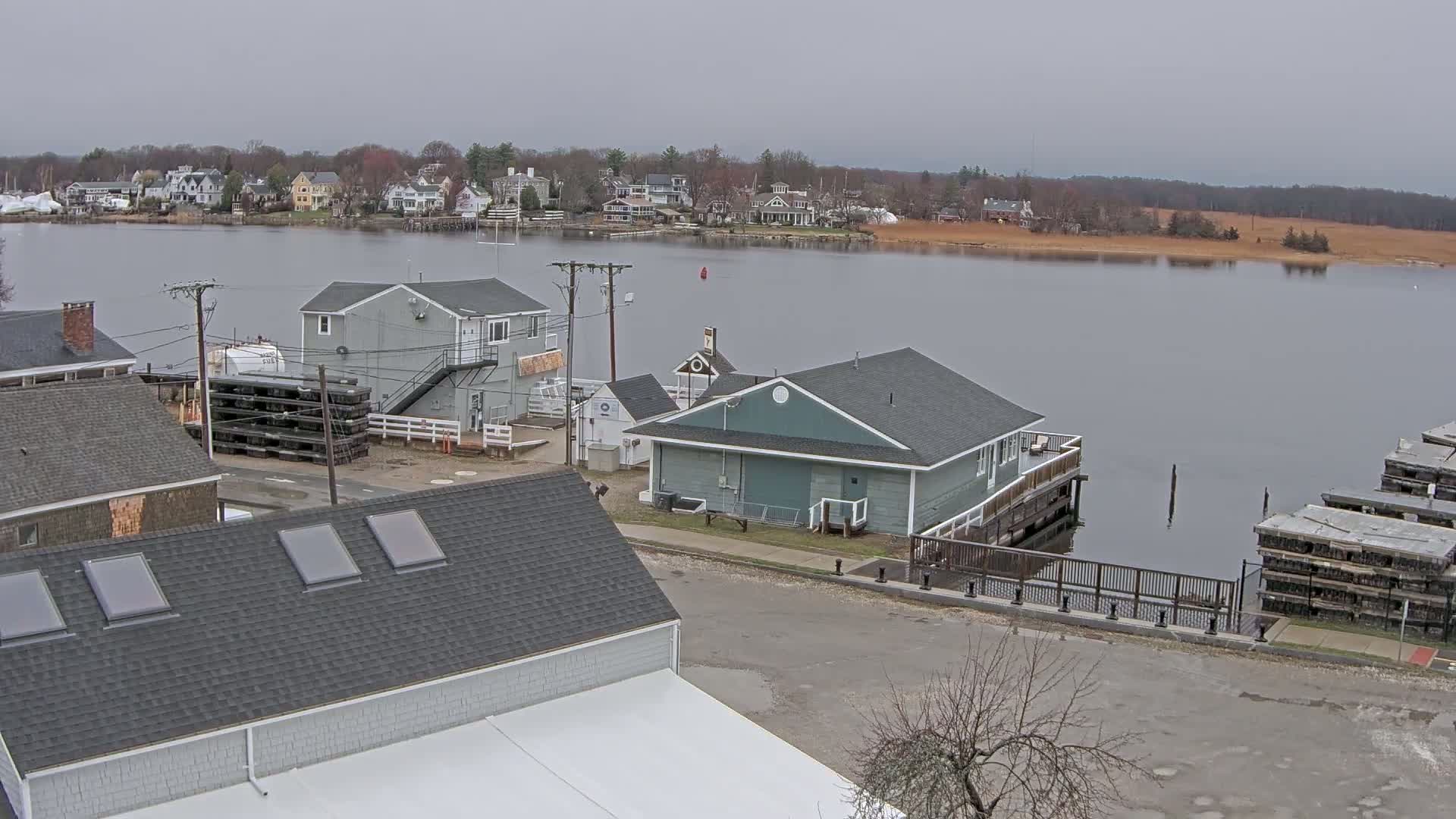 An overcast day reveals a waterfront scene with several buildings, including a teal-colored structure near a calm body of water, and stacks of what appears to be lumber or building materials near the shore.