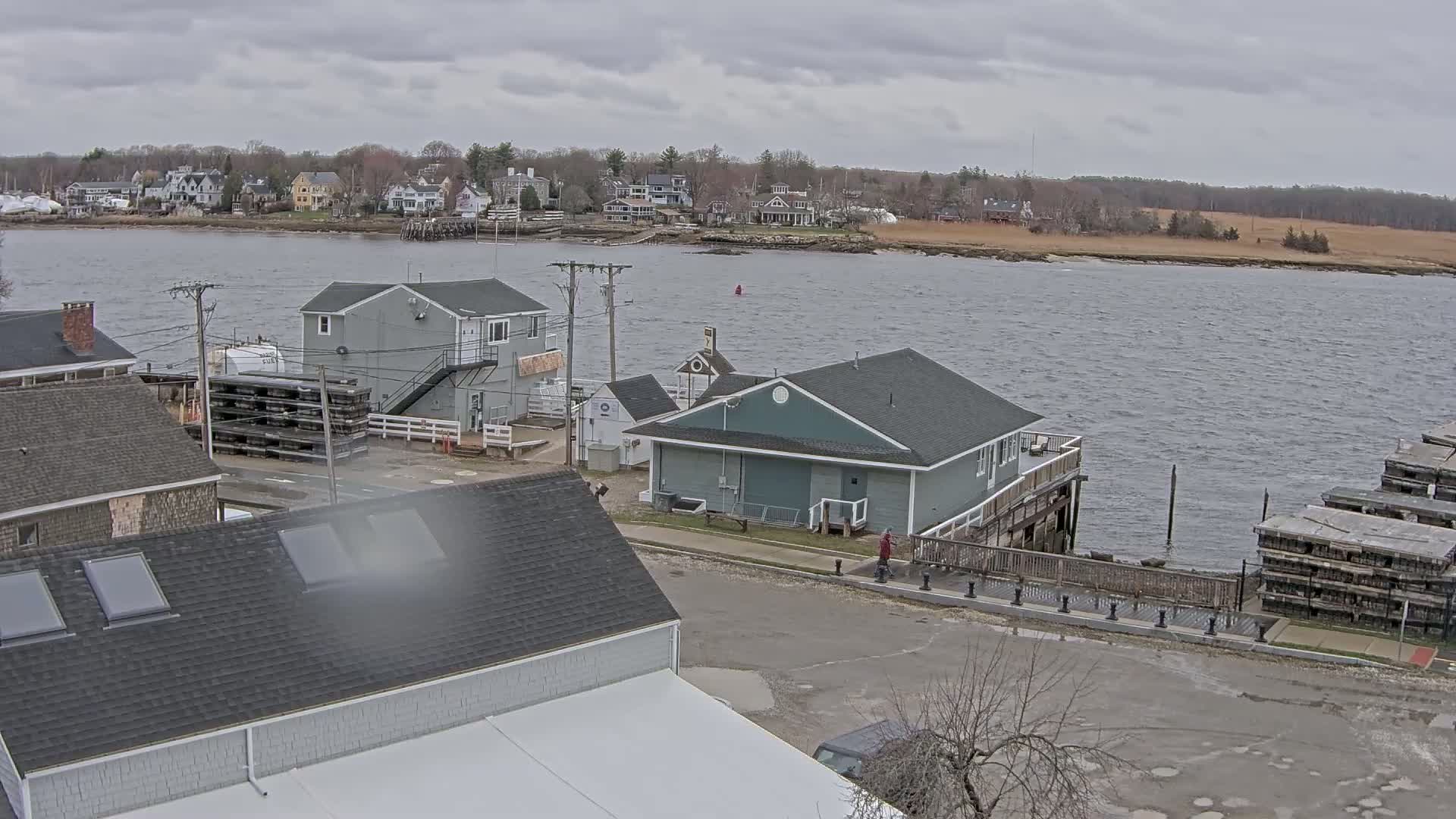 A waterfront view of a town on an overcast day shows several buildings, a calm body of water, and stacks of what appear to be lobster traps.