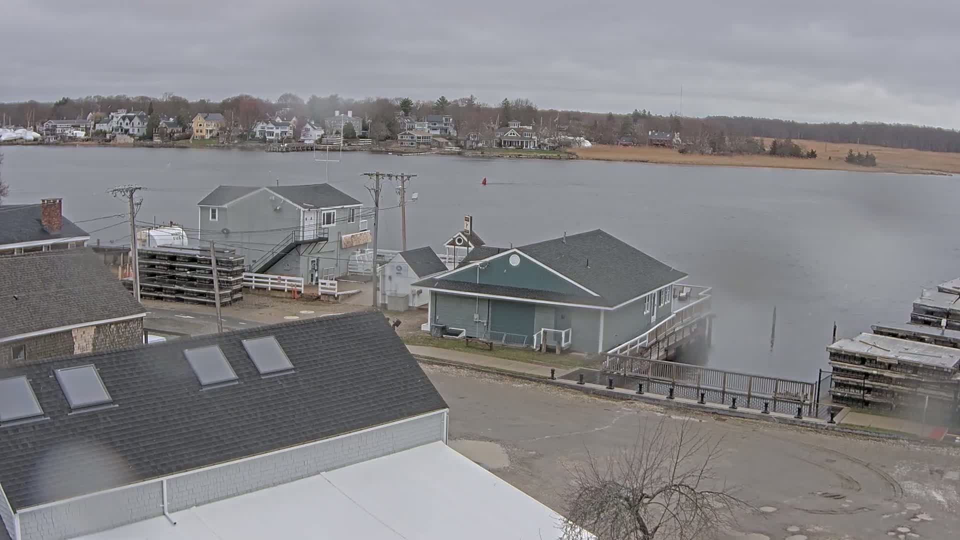 A waterfront view of several buildings, including a gray two-story building and a teal single-story building near a calm body of water under an overcast sky.