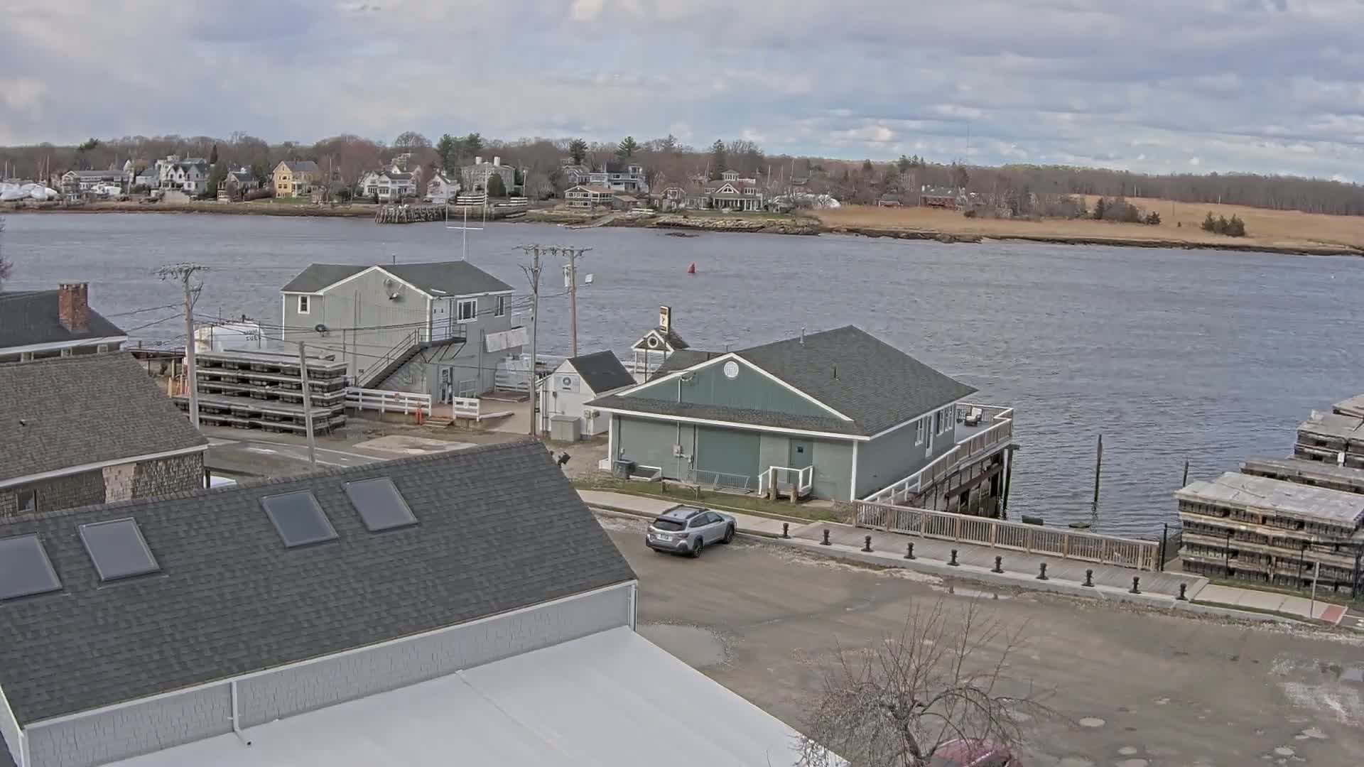 A waterfront scene on an overcast day shows several buildings near a calm river with a town visible across the water.