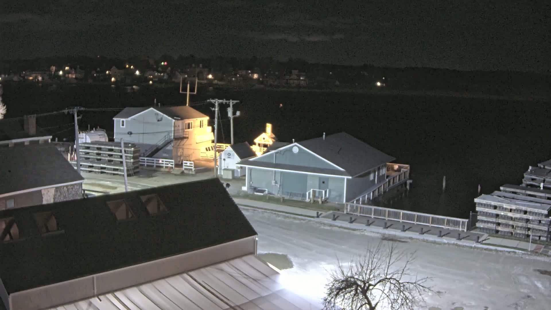 A nighttime view of a waterfront town shows several buildings near a body of water, with snow or ice visible on the ground and stacks of what appear to be oyster cages nearby.