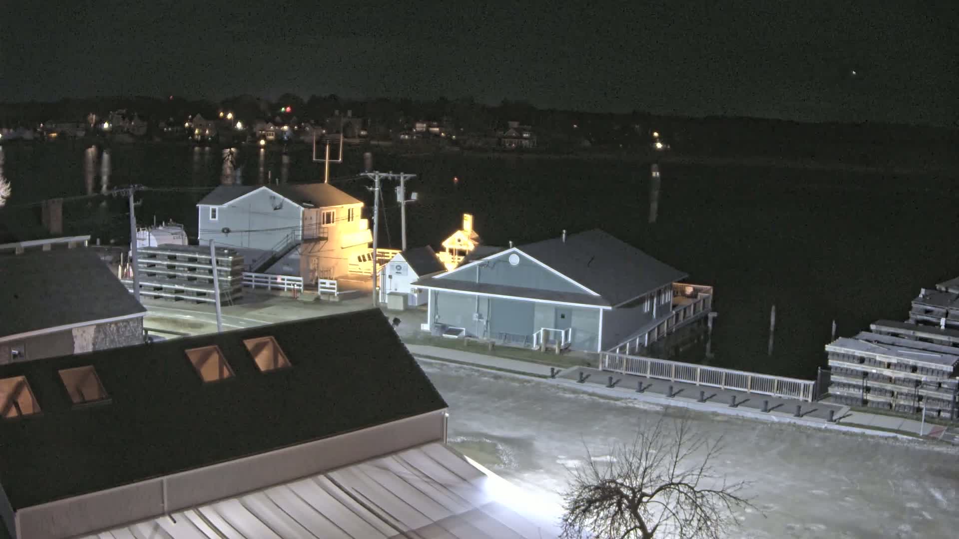 A nighttime view of a waterfront area with several buildings, a calm body of water, and stacks of wooden pallets.