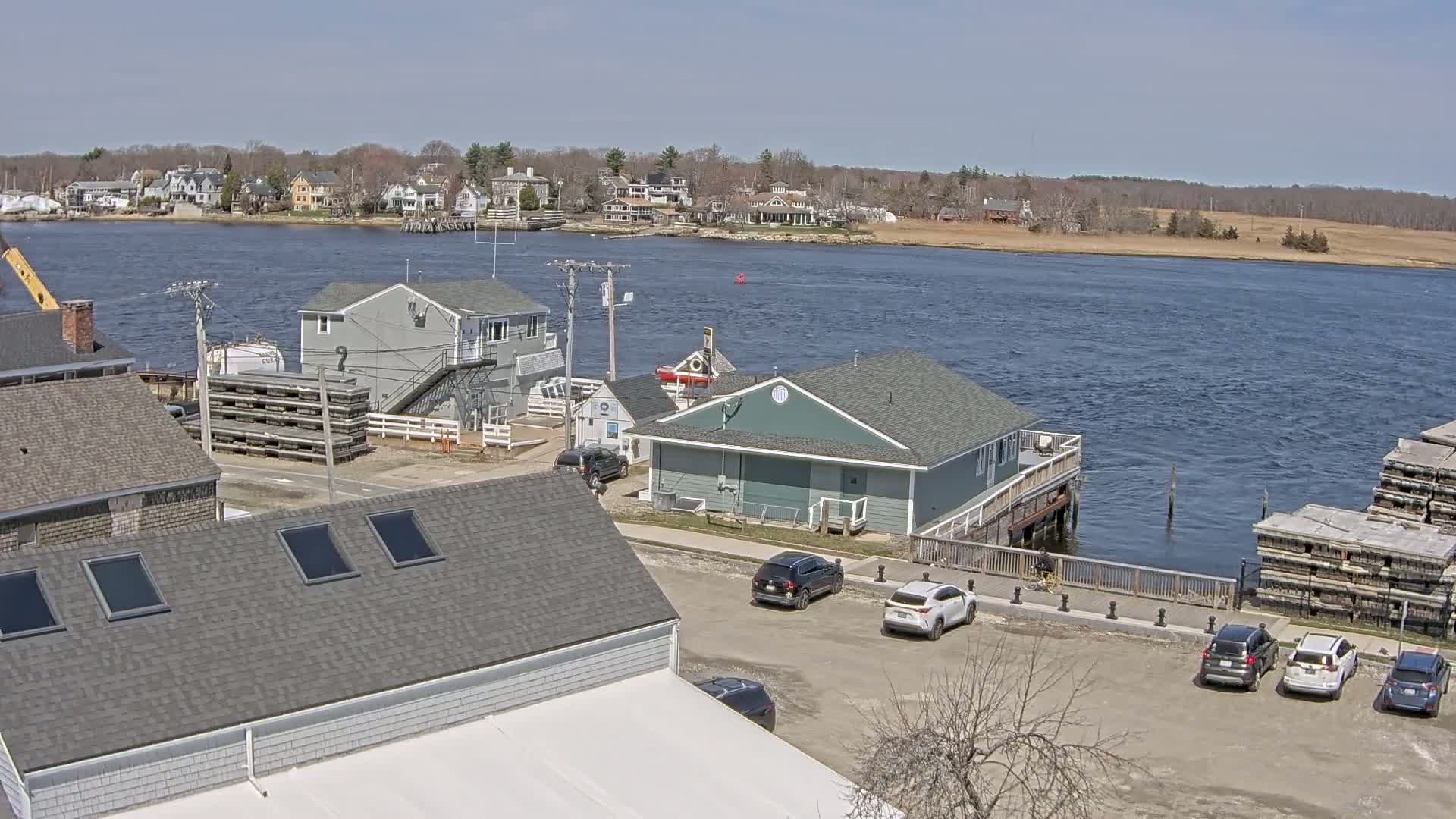 A waterfront view of a small town on a sunny day shows several buildings near a calm river, with cars parked in a lot and stacks of what appear to be wooden materials by the water's edge.