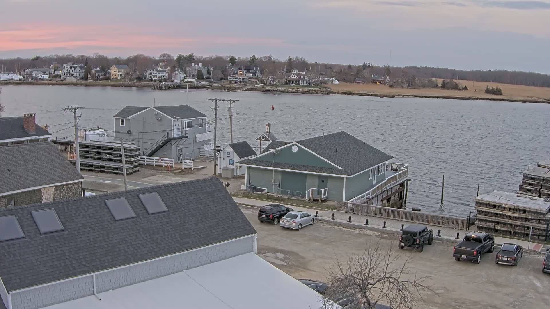 A waterfront scene at dusk shows several buildings, parked cars, and stacks of what appear to be oyster cages along a calm body of water, with a residential area visible in the distance.