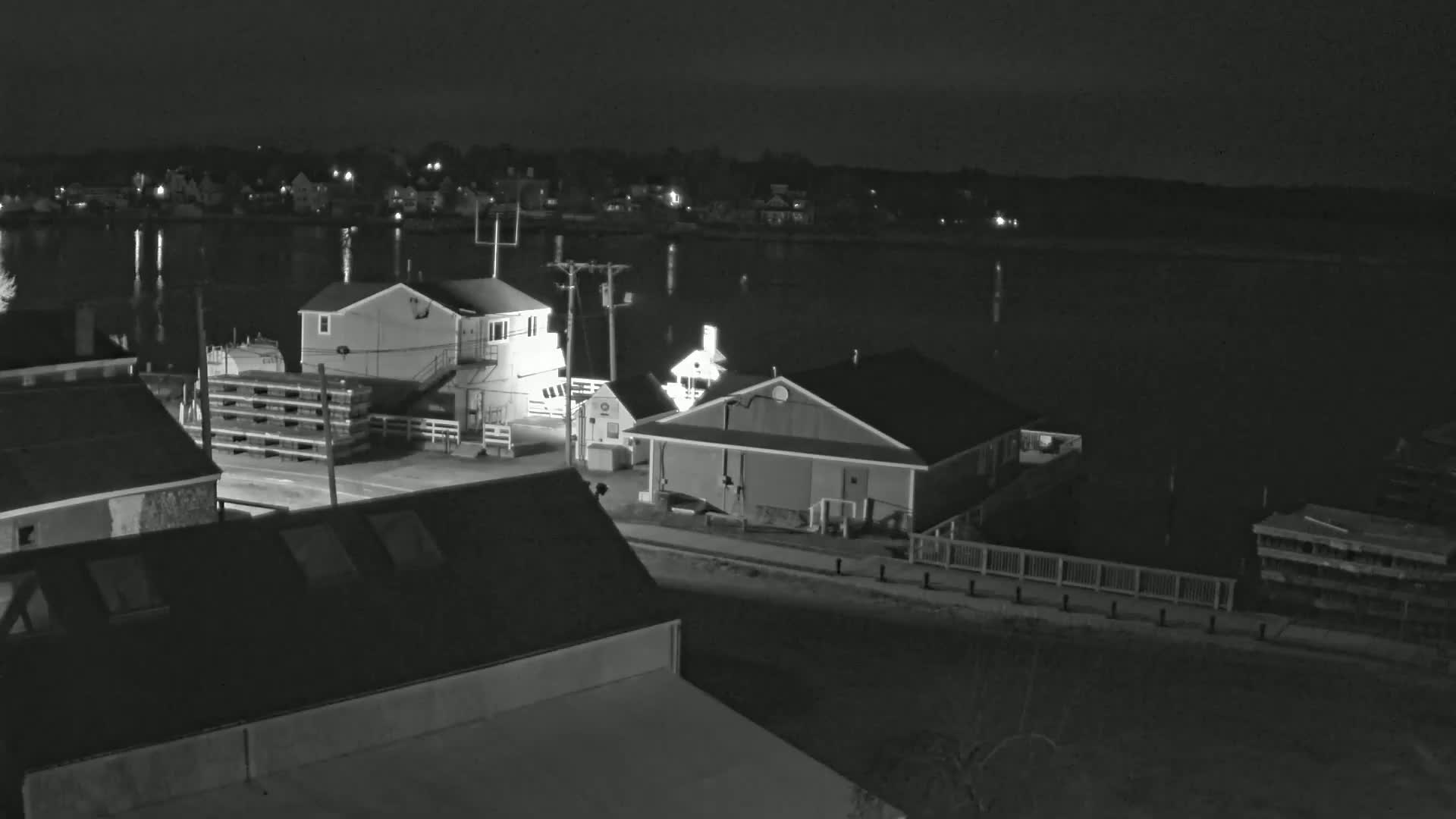 A nighttime, high-angle view of a waterfront area with several buildings, a calm body of water, and stacked wooden pallets.