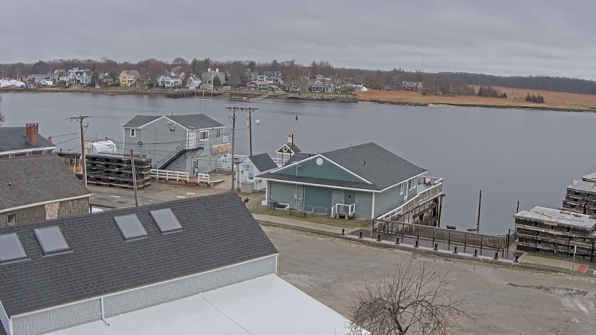 A waterfront scene on an overcast day shows several buildings near a calm body of water, with a small town visible in the background.