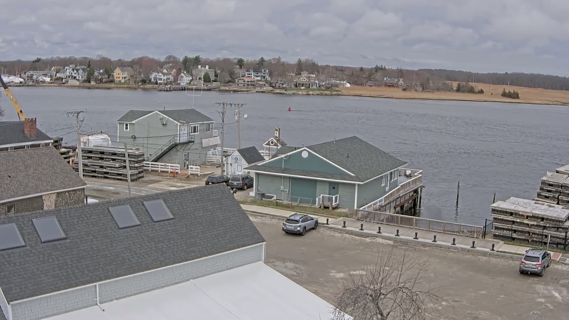 An overcast day reveals a waterfront scene with several buildings, cars parked in a lot, and stacks of wooden planks near a calm body of water, beyond which lies a residential area.