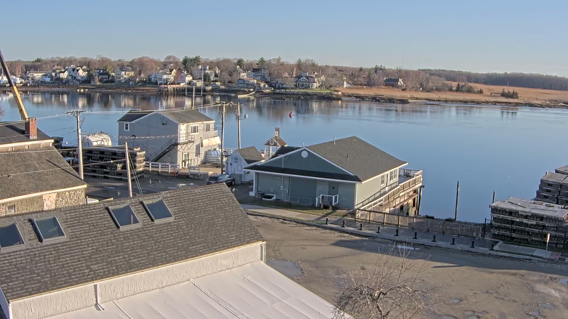 A waterfront village scene on a sunny day shows several buildings, a calm river, and stacks of what appear to be lobster traps.