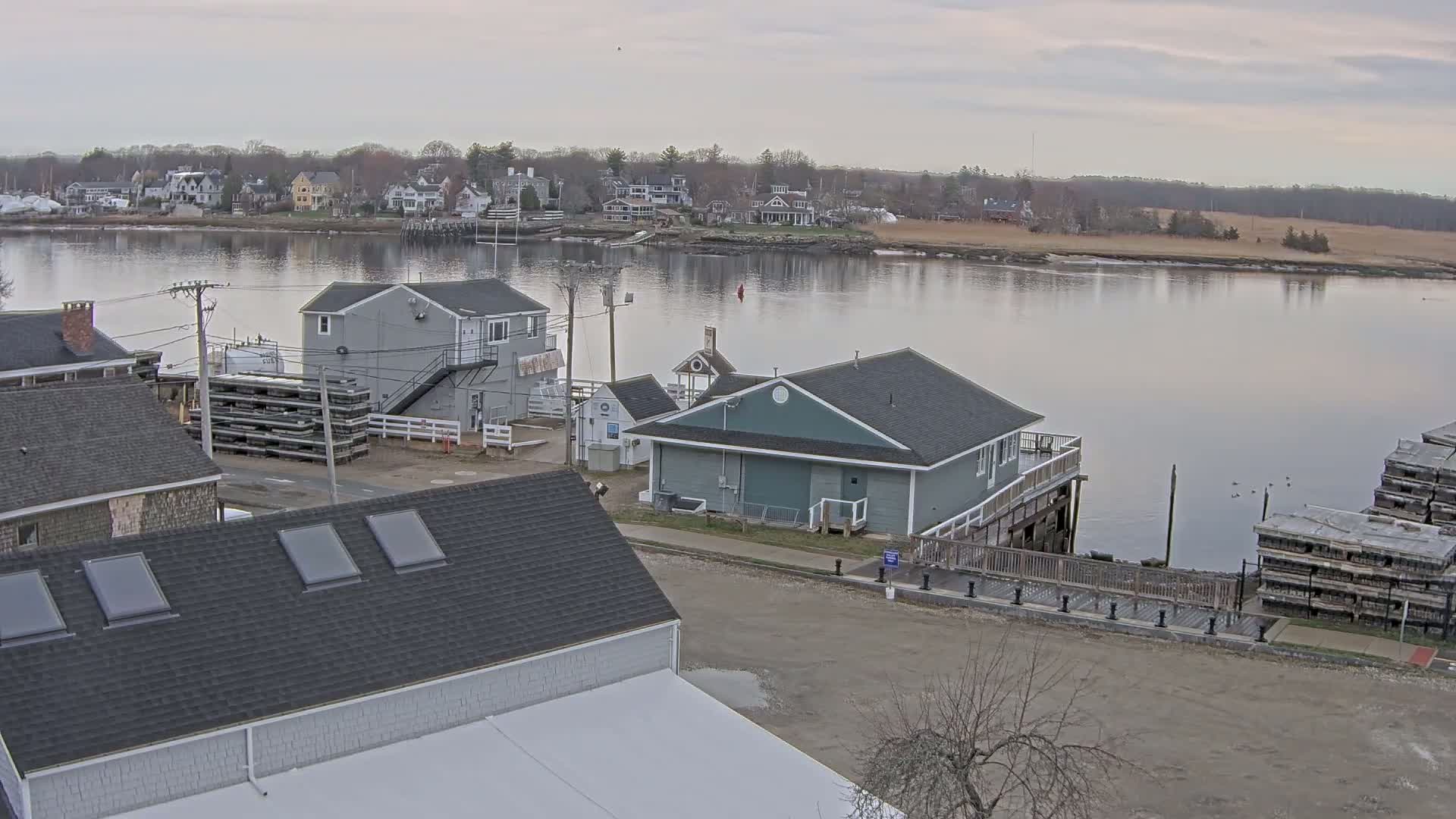 A calm river reflects a cloudy sky above a waterfront scene of houses, small buildings, and stacked wooden structures.