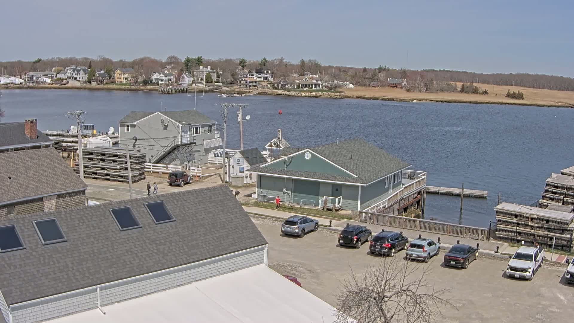 A waterfront scene on a sunny day shows a small town with houses and buildings along a calm river, several cars parked in a lot, and some stacked wooden crates by the water.