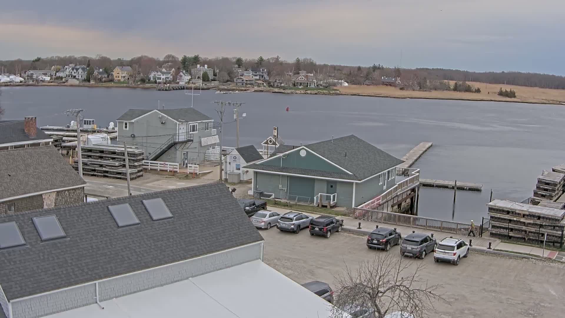 A waterfront scene on an overcast day shows several buildings, cars parked in a lot, and a calm body of water with docks and boats in the distance.