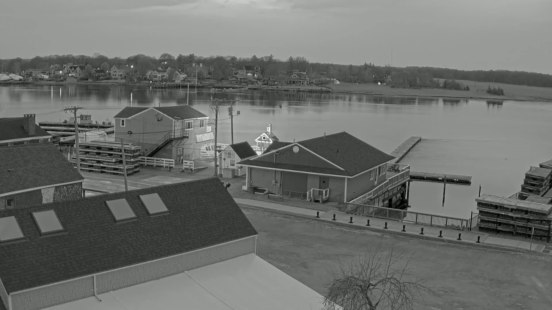 A grayscale view of a waterfront town at dusk, showing several buildings near a calm river with docked boats and stacks of wooden crates.