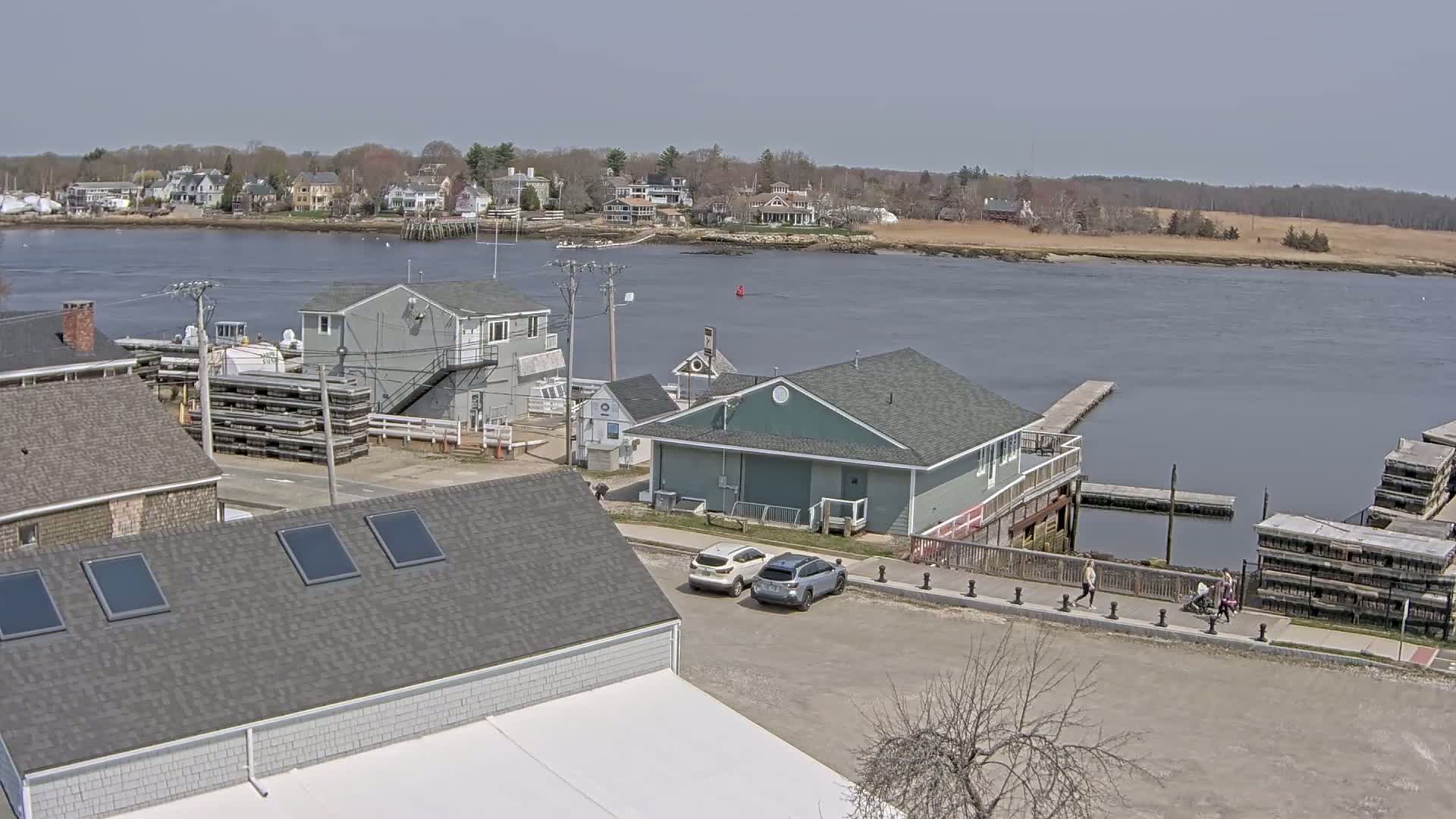 A waterfront scene on a partly sunny day shows several buildings, a parking lot with two cars, and people walking along a boardwalk next to a calm body of water with docks and boats.
