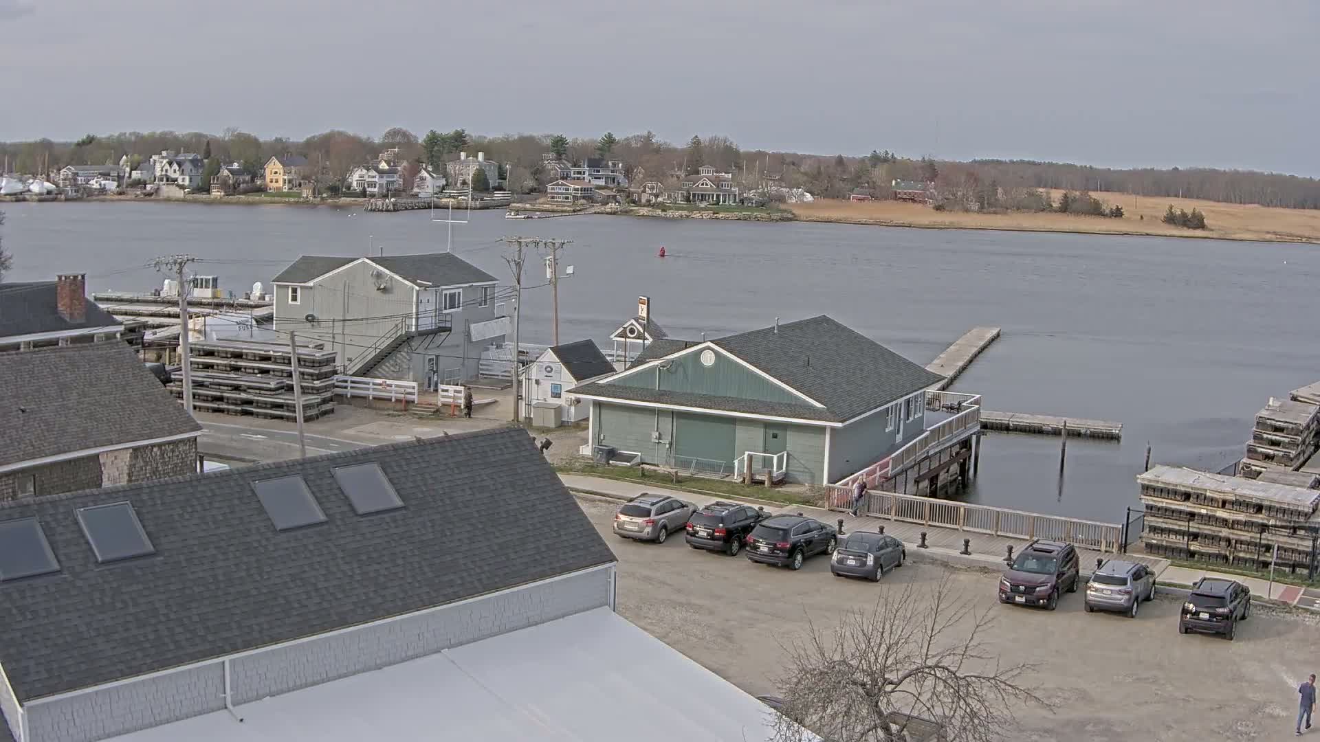 A waterfront scene on an overcast day shows several buildings, parked cars, and boats, with a calm body of water in the background.