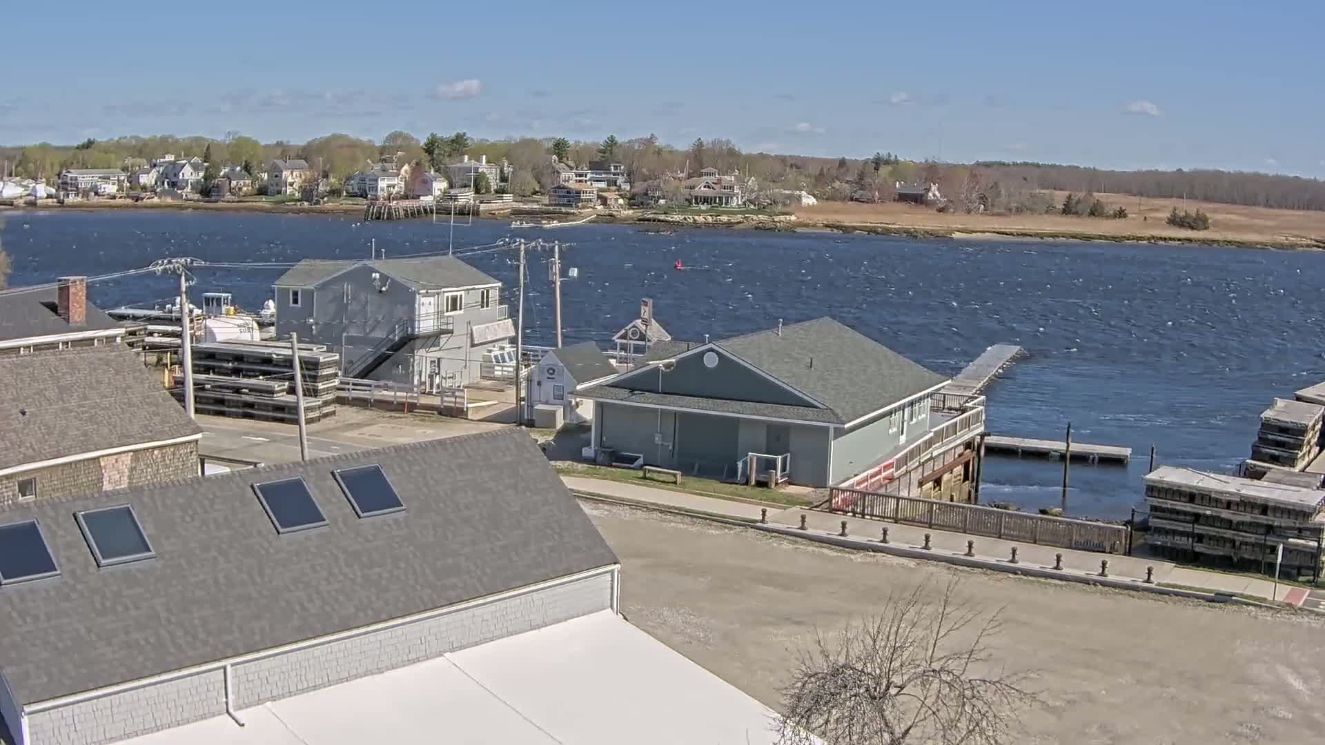 A waterfront scene on a windy day shows several gray buildings near a body of water with small waves, across from a residential area with houses and trees.