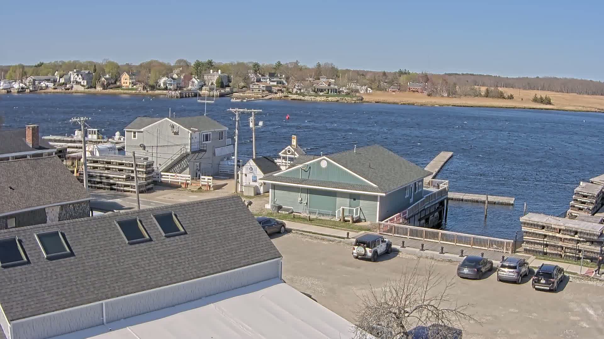 A waterfront scene on a sunny day shows several buildings, cars parked in a lot, a small boat dock, and a body of water with small waves.