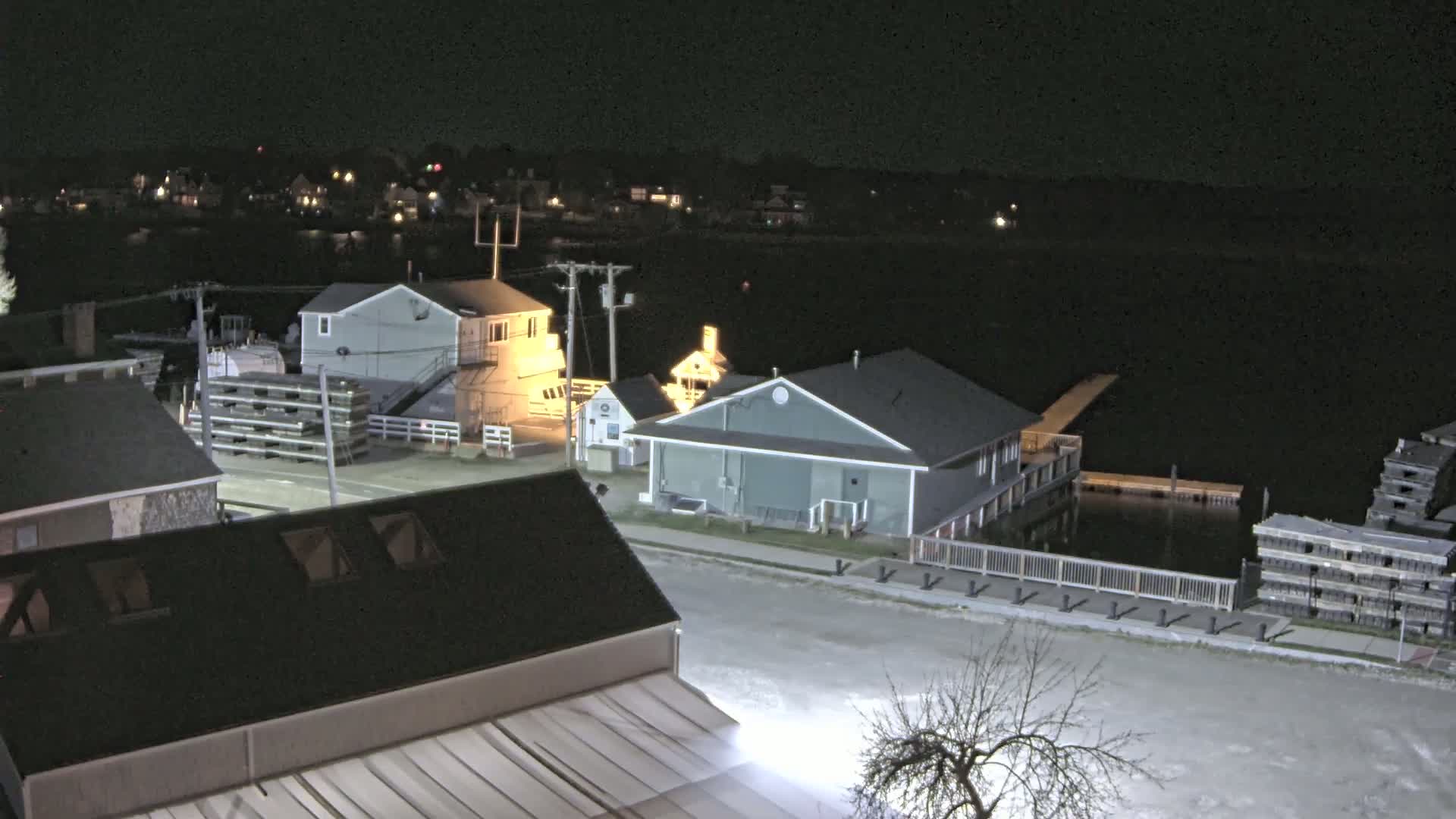 At night, a waterfront area shows several buildings near a dark body of water, with stacks of what appear to be lobster traps near a dock.