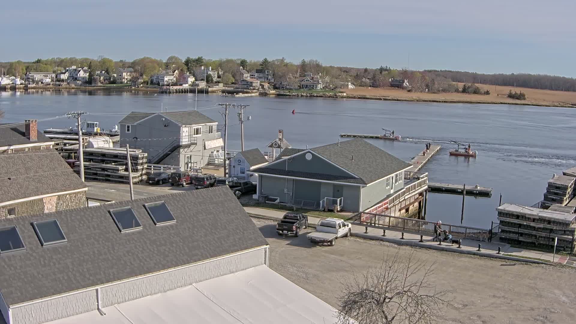 An aerial view of a waterfront town on a sunny day shows several buildings, parked cars, a small marina with boats, and a person in a red kayak on the water.