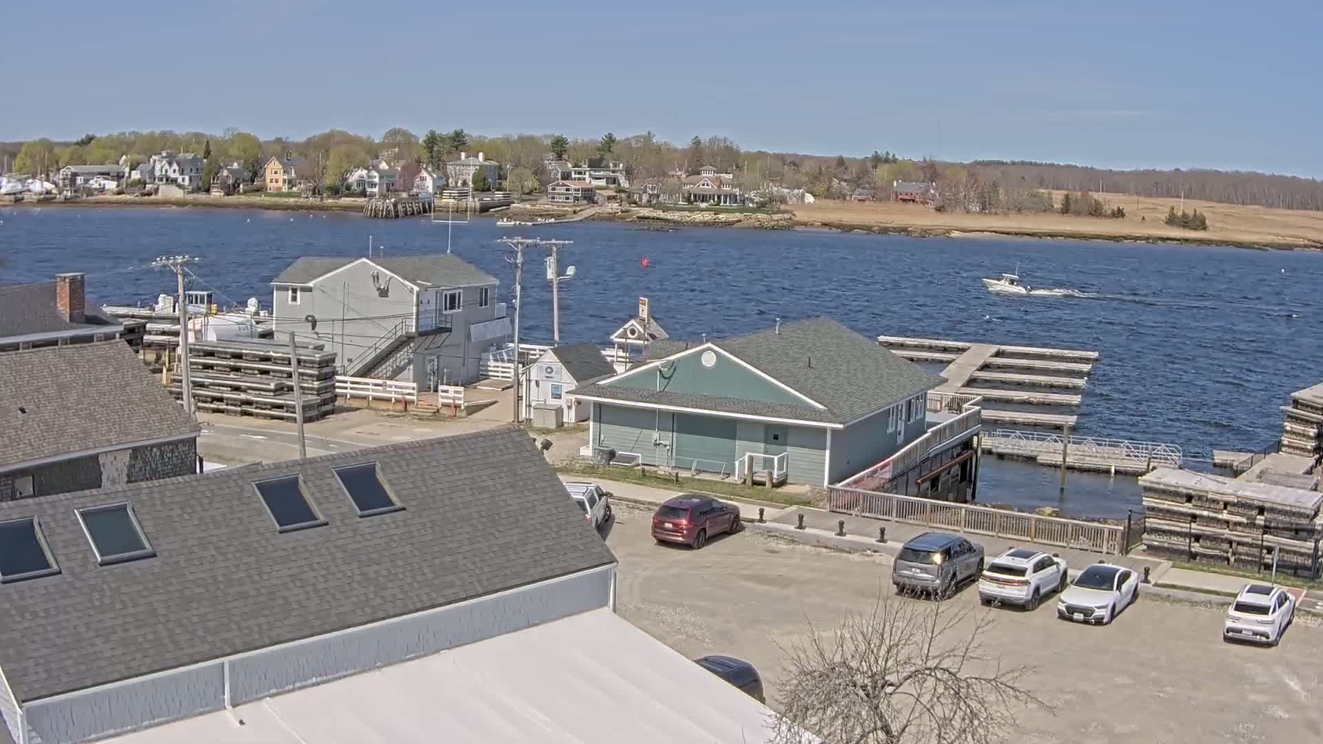 A waterfront town on a sunny day with a few boats on the water, several buildings, and cars parked in a lot near a marina.
