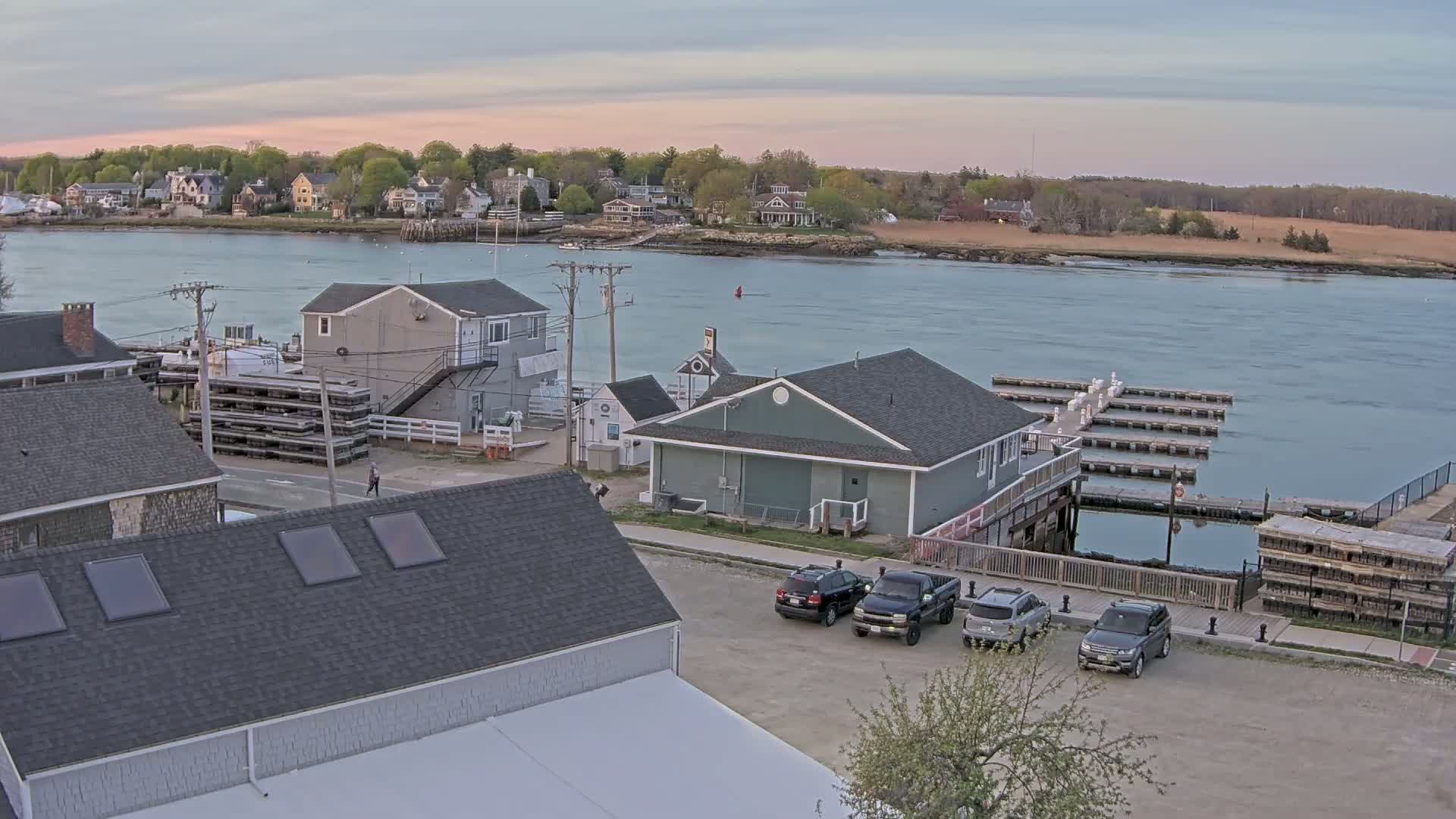 A calm, twilight view of a waterfront town with several buildings, a parking lot containing several vehicles, a marina, and a body of water extending to a tree-lined shore in the background.