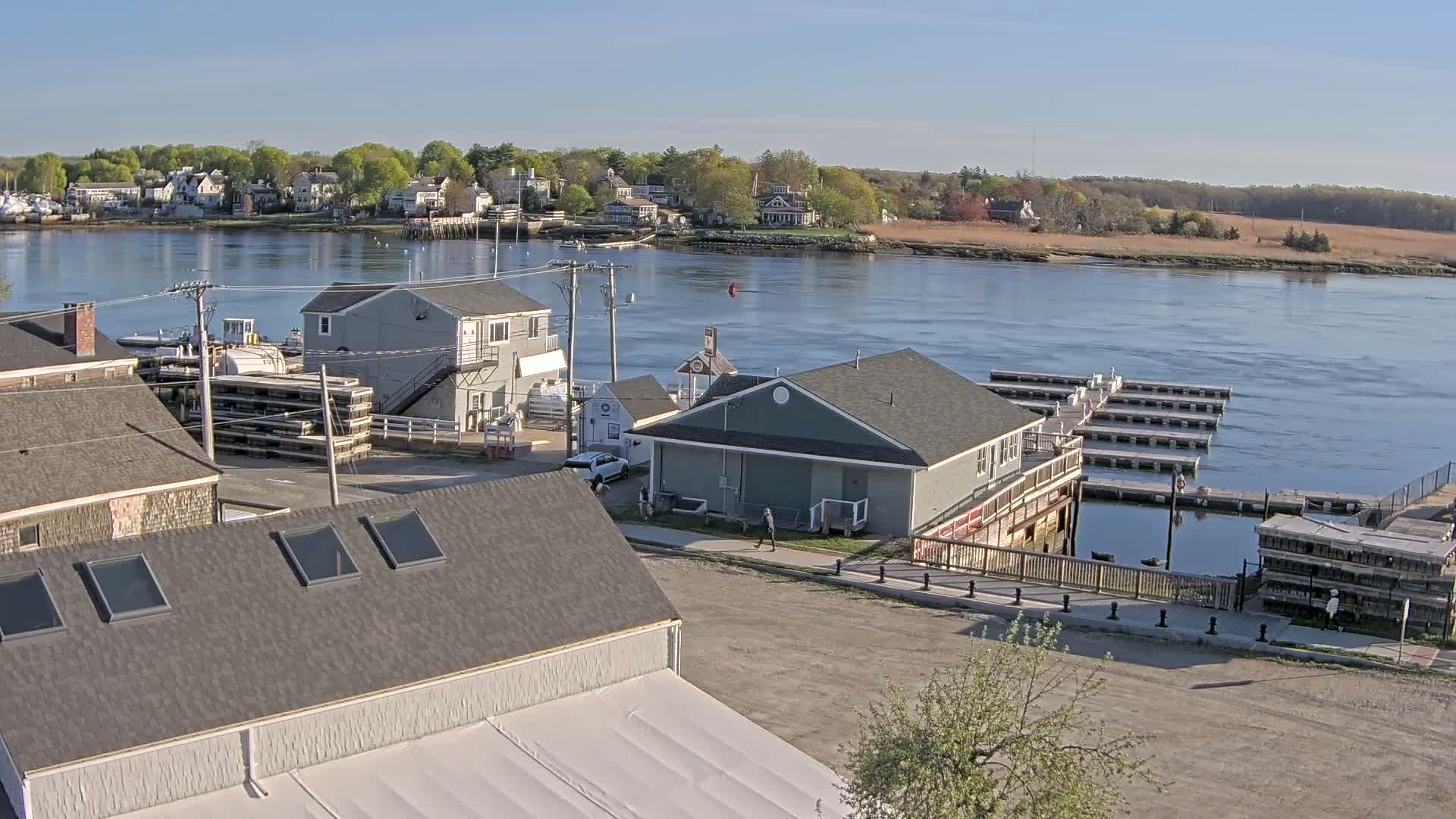 A waterfront scene on a sunny day shows a village with several buildings, including a gray two-story building near a body of water with numerous docks and a few boats.