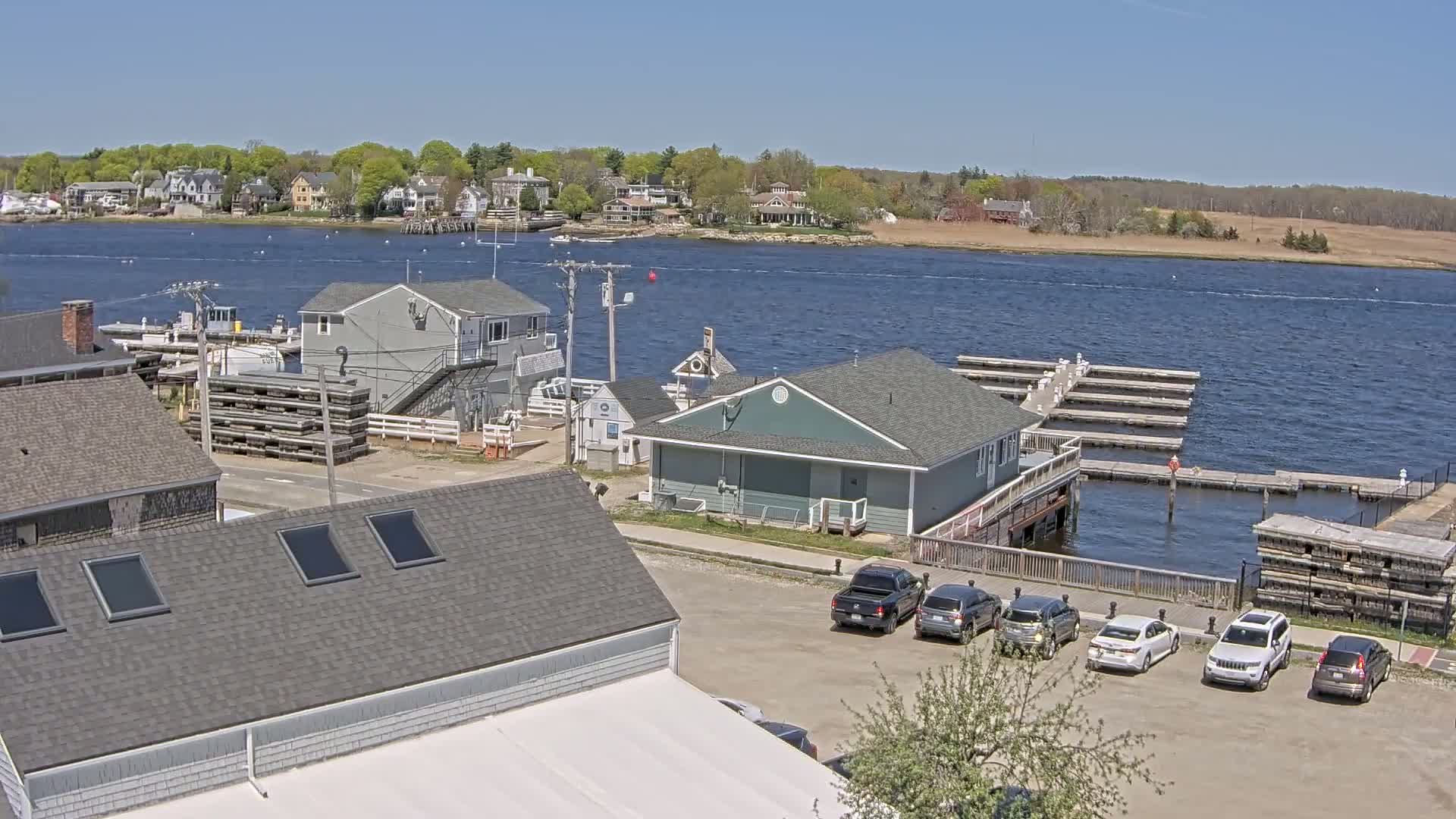 A waterfront scene on a sunny day shows a small town with houses, a marina with boats docked, and several cars parked in a lot next to a building.