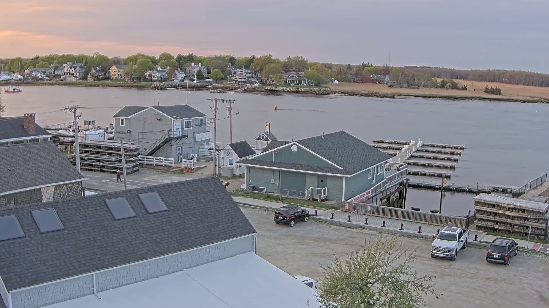 A waterfront town with several buildings, a parking lot with cars, and a marina with docks under a partly cloudy sky.