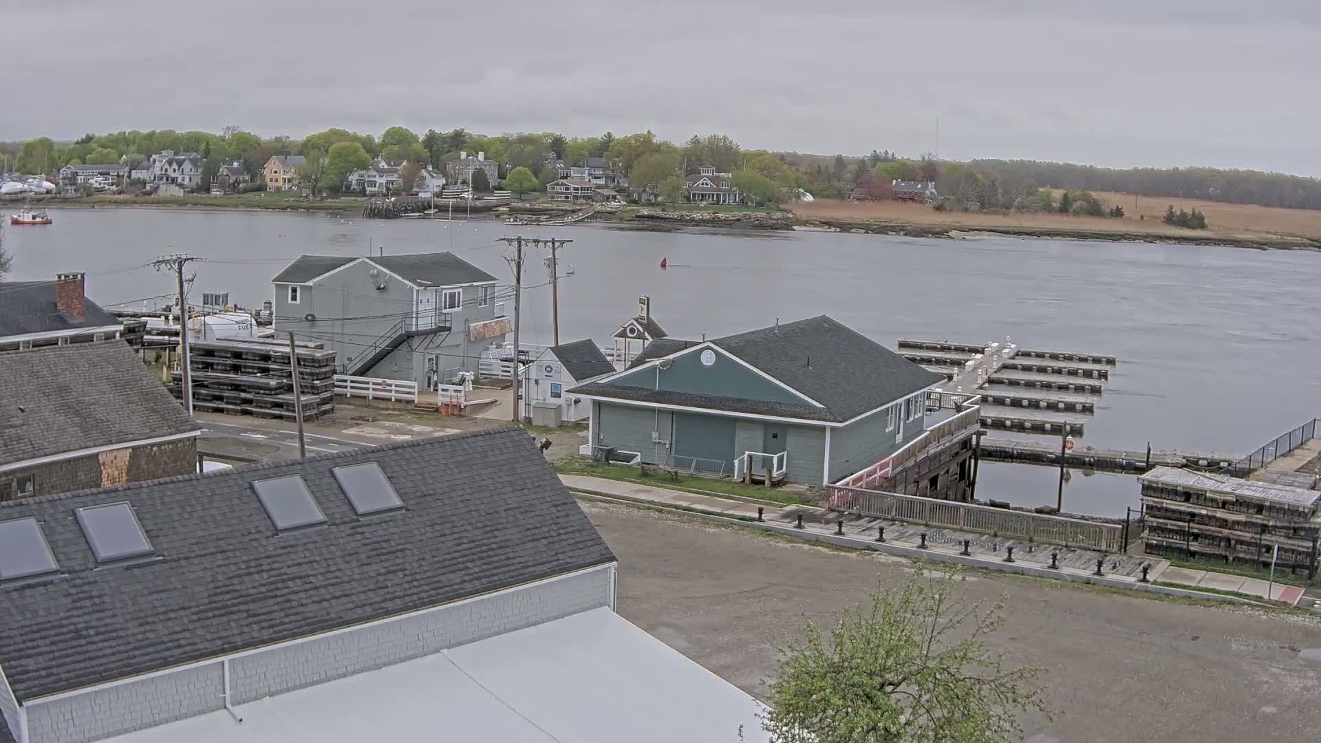 A waterfront scene on an overcast day shows several buildings near a marina with boat slips and a calm river in the background, across which lies a line of houses and trees.