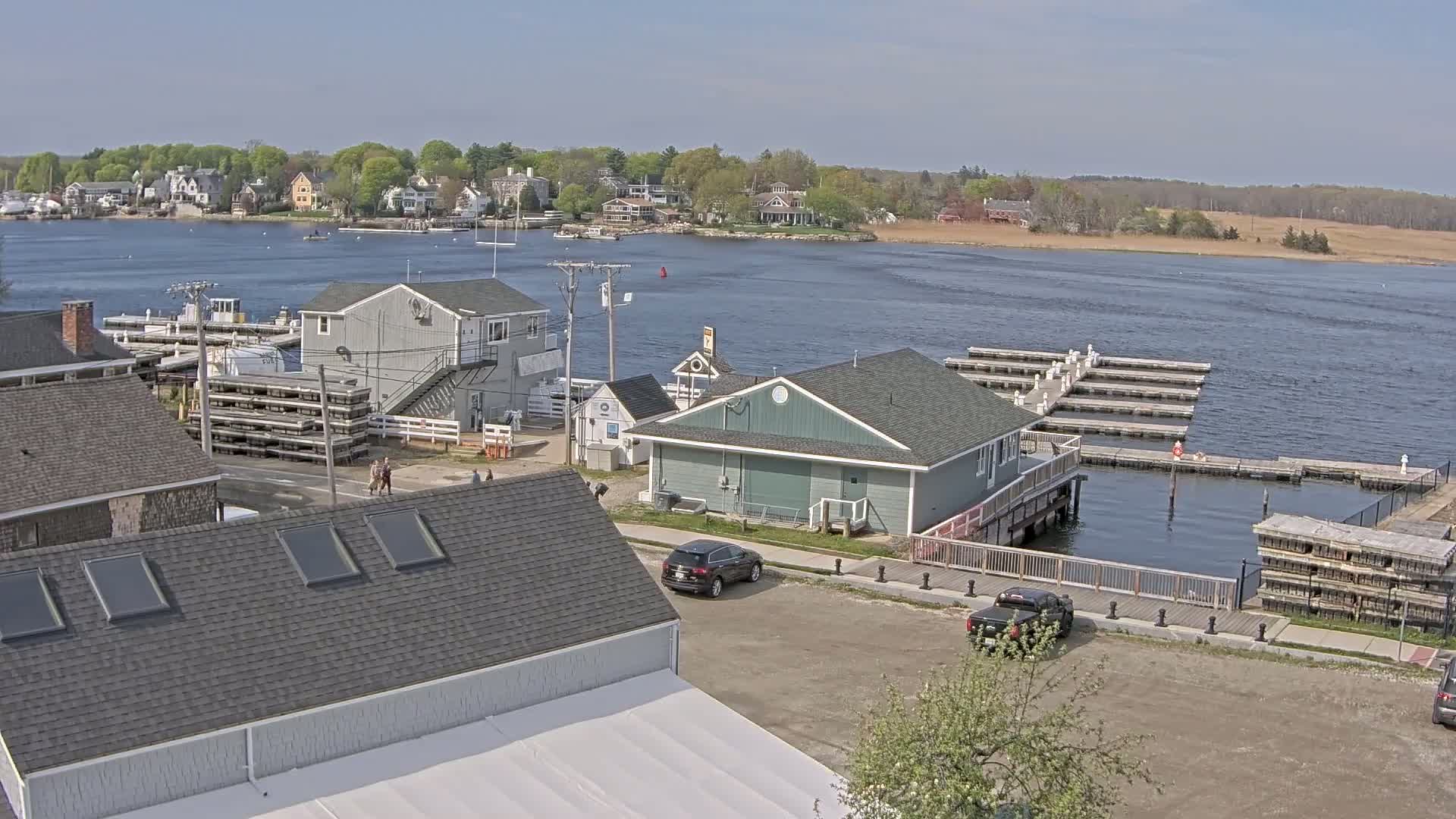 A waterfront town on a sunny day shows several buildings near a harbor with boats and docks, and cars parked in a lot.