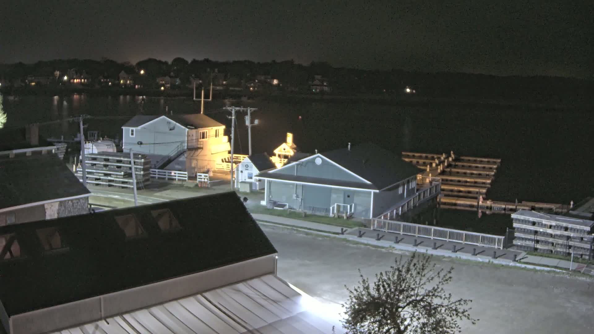 A nighttime view of a waterfront town with several buildings illuminated near a calm body of water, featuring a marina with docked boats.