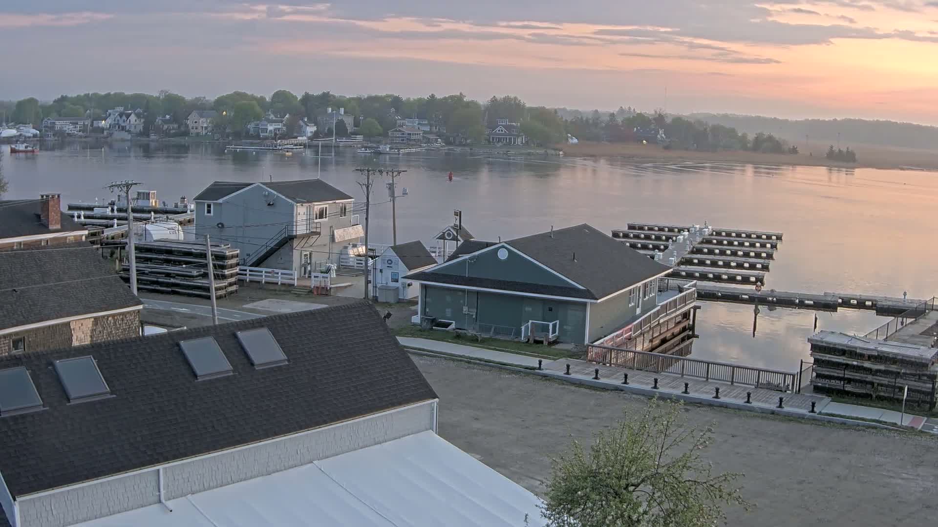 A waterfront town at sunrise, with calm water reflecting the pastel sky, features several buildings, docks, and stacked materials near a harbor.