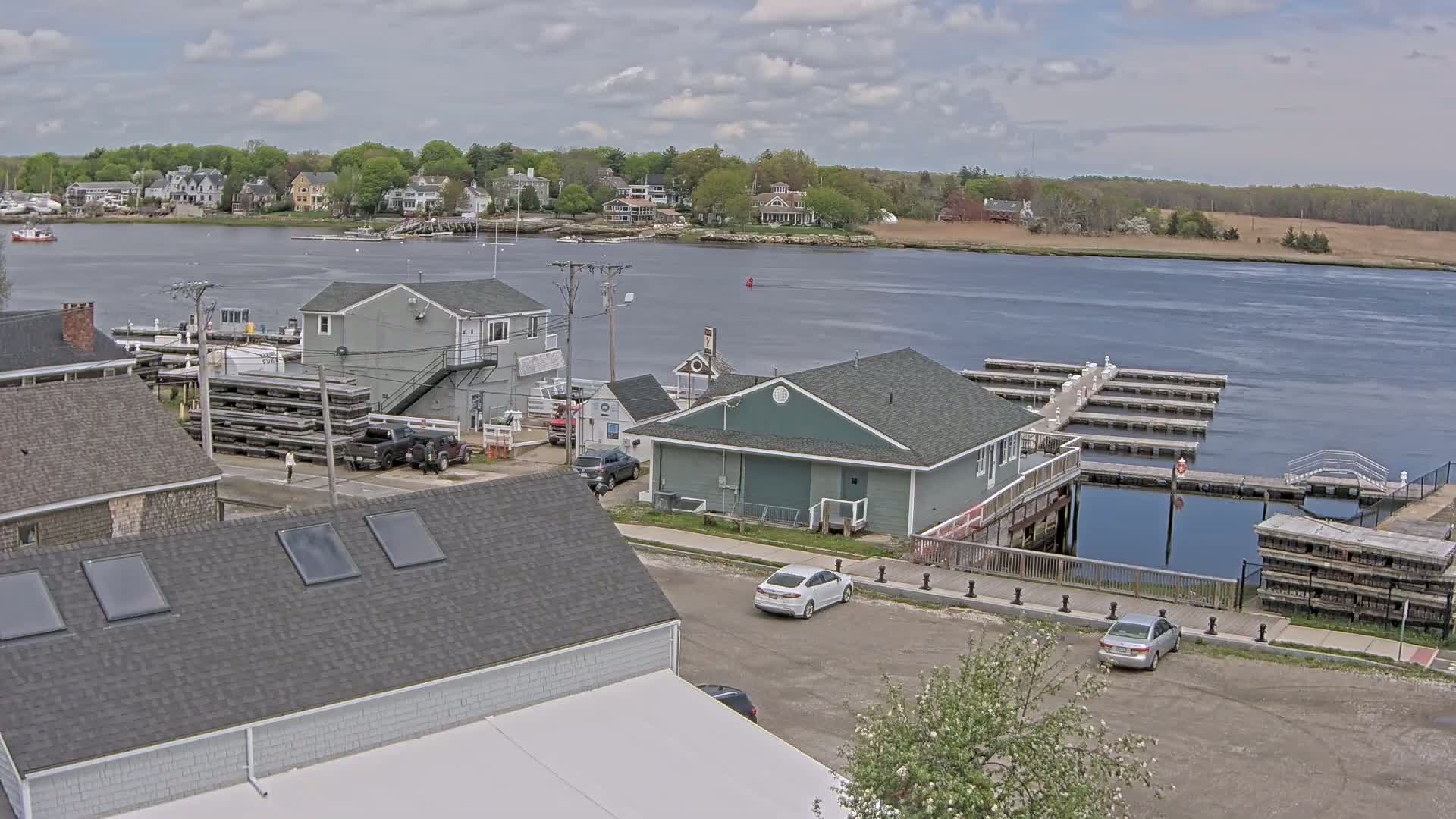An overcast day reveals a waterfront scene with a marina, several buildings, parked cars, and a river leading to houses on the opposite shore.