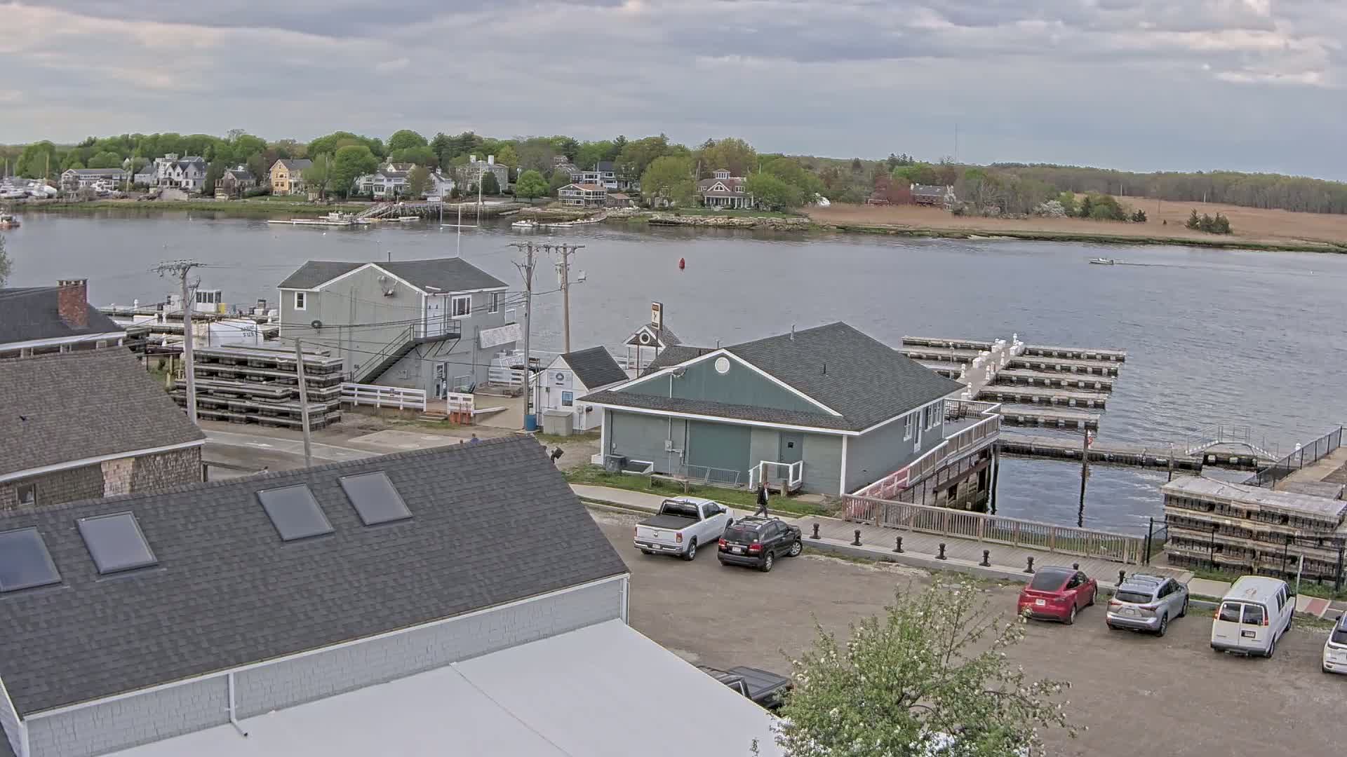 A waterfront scene on an overcast day shows a small town with houses along a river, a marina with boats and docks, and a parking lot with several vehicles.