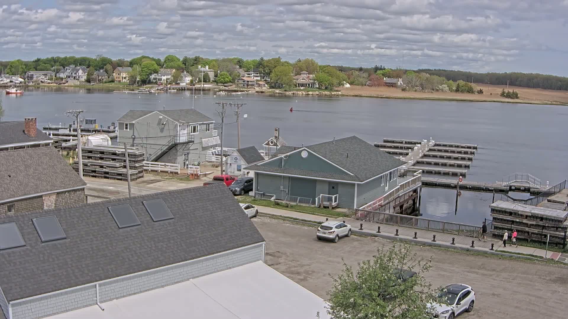 A waterfront town on a partly cloudy day features several buildings, a parking lot with cars, a boat dock, and a calm river with houses visible across it.