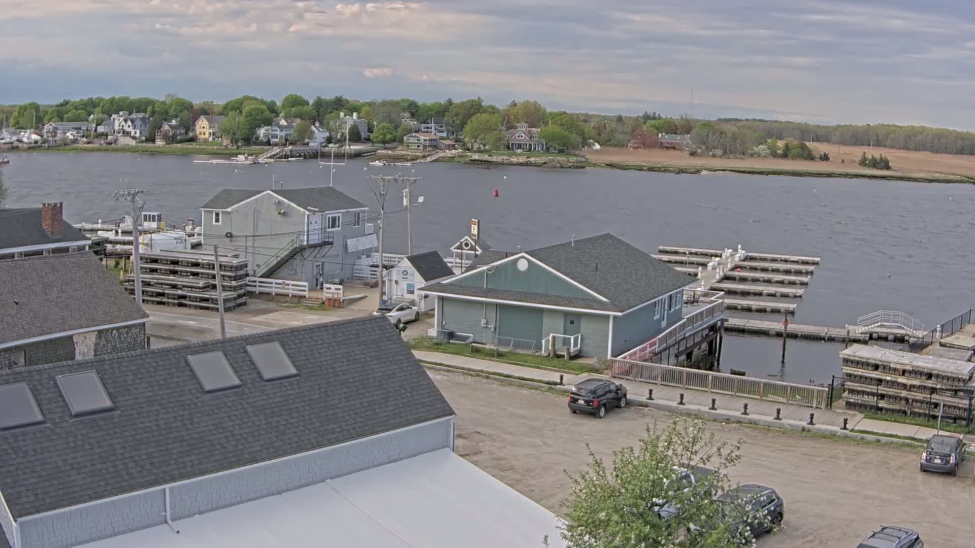 An overcast day shows a waterfront view of a small town with buildings, docked boats, and cars parked near a body of water.