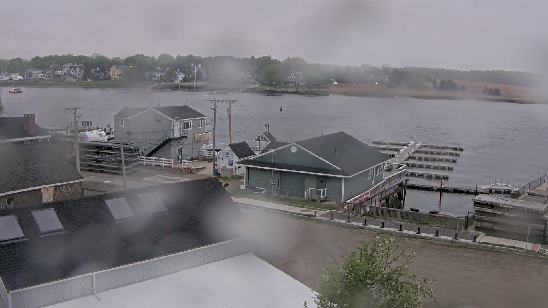 A rain-blurred view shows a waterfront area with several buildings, a boat dock, and a calm body of water under an overcast sky.