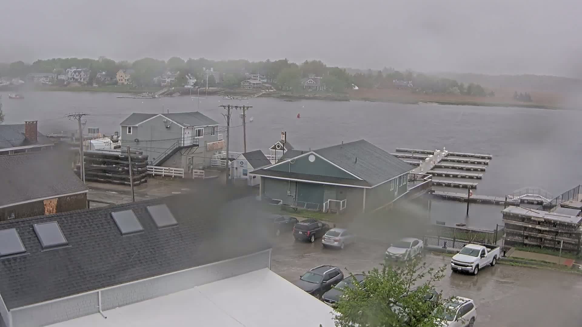 A rain-soaked coastal town is viewed from above, showing buildings, a parking lot with cars, a marina with docks, and a body of water in the distance.
