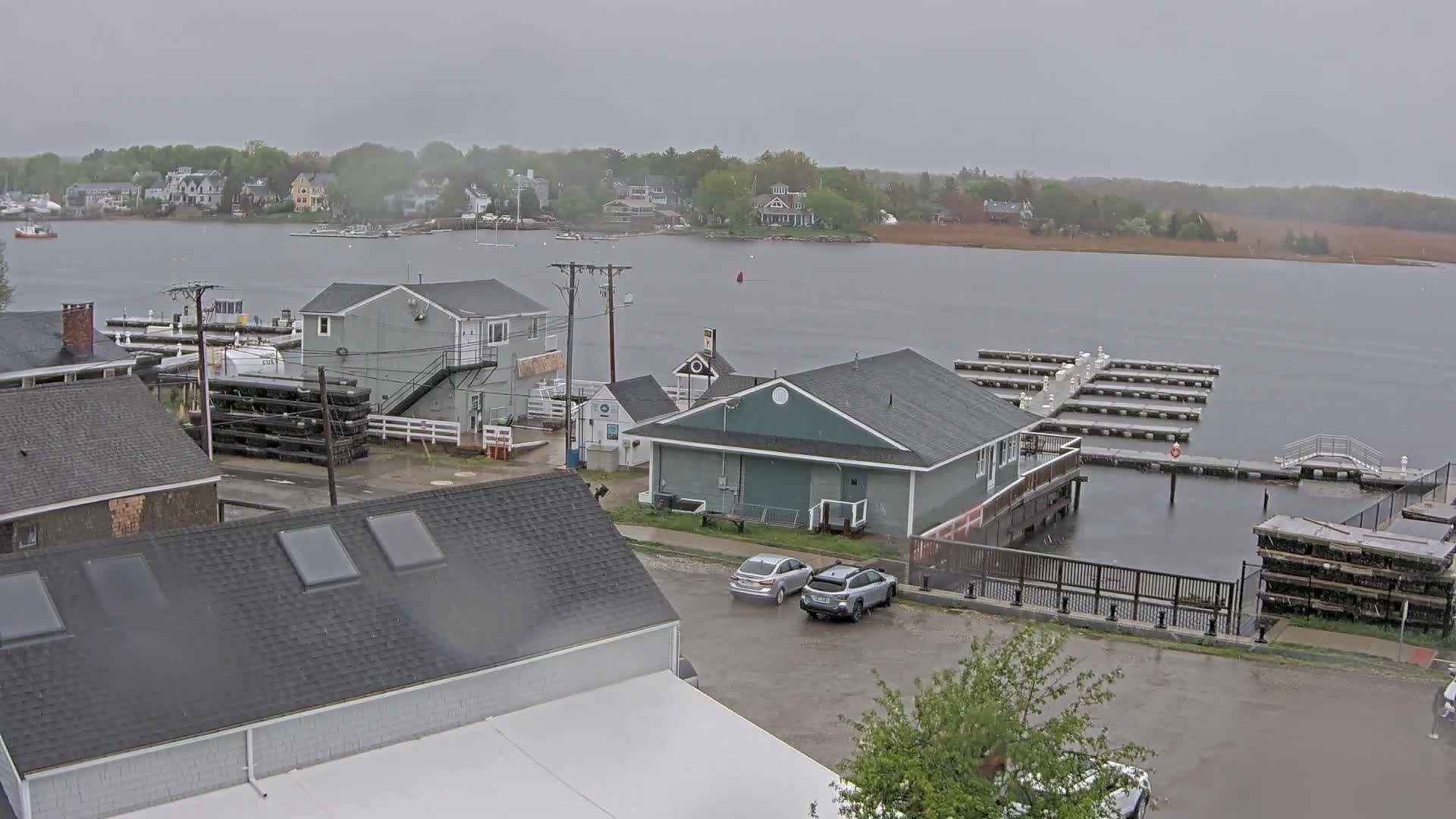 A rain-swept waterfront scene shows a marina with boats docked, several buildings, and a few cars parked in a lot.