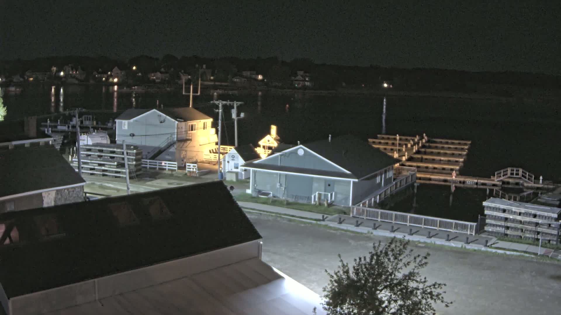A nighttime view of a waterfront town with several buildings illuminated along a calm body of water, featuring docks and a small amount of land-based activity.