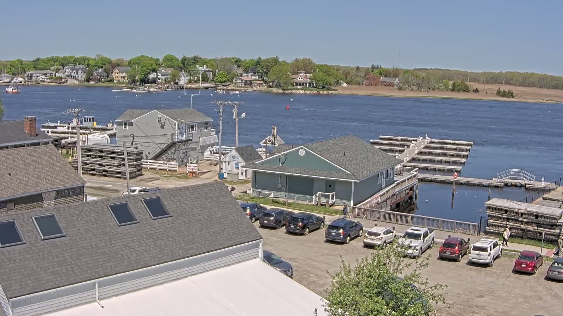 A waterfront scene on a sunny day shows a harbor with boats docked at piers, several buildings, and a parking lot with cars.