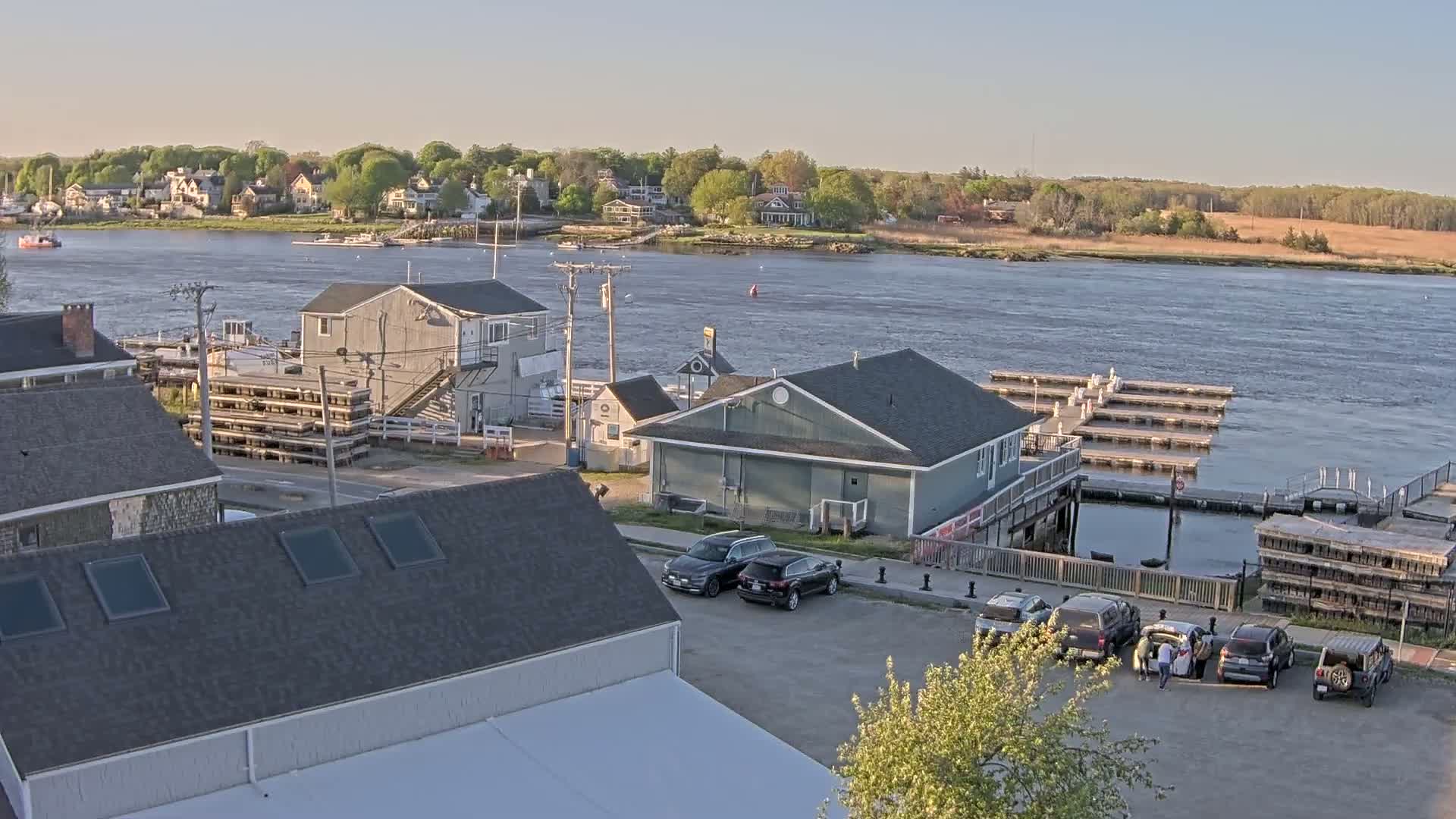 A waterfront scene on a calm, sunny day shows a harbor with several buildings, docked boats, and cars parked in a lot, with a village visible across the water.