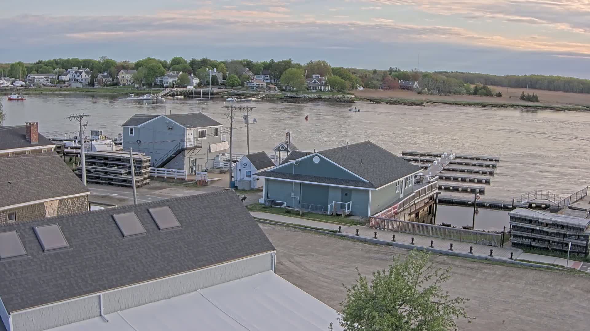 A waterfront town with several buildings, docks, and boats on a calm river under a cloudy sky.