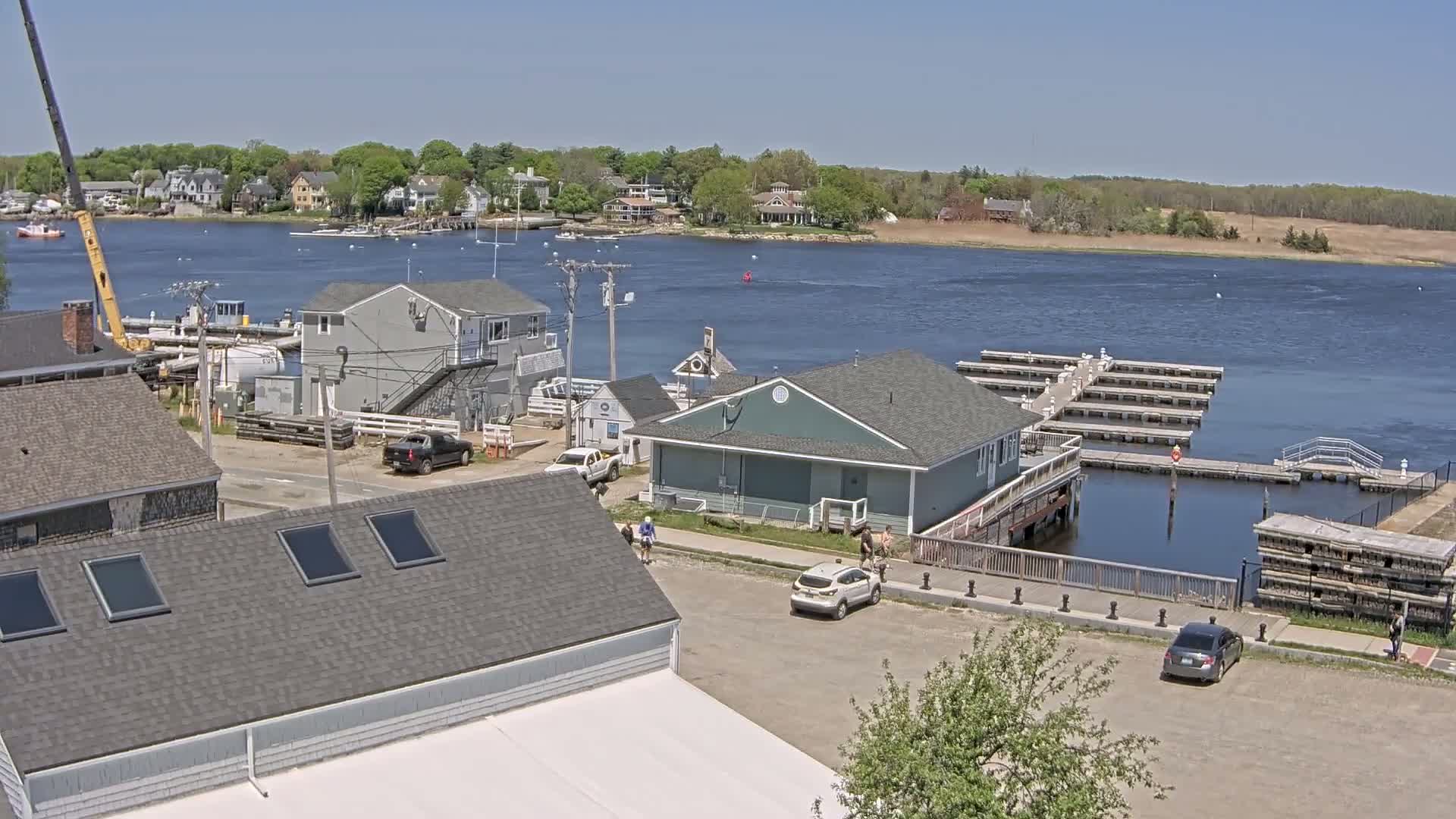 A sunny day overlooks a waterfront area with buildings, a parking lot, a marina with docks, and a calm river.