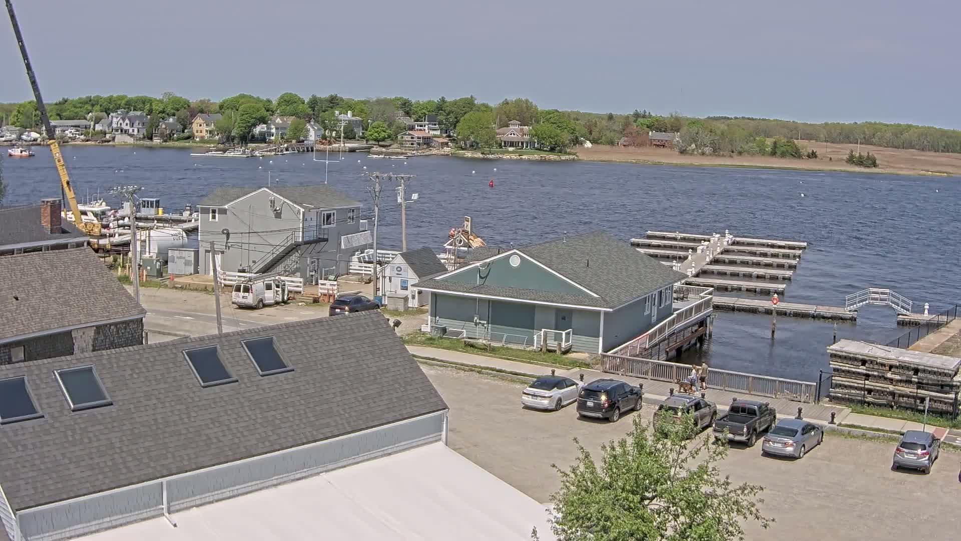 A waterfront scene on a sunny day shows a marina with boats, several buildings, and cars parked in a lot, with green trees and houses visible in the background across the water.