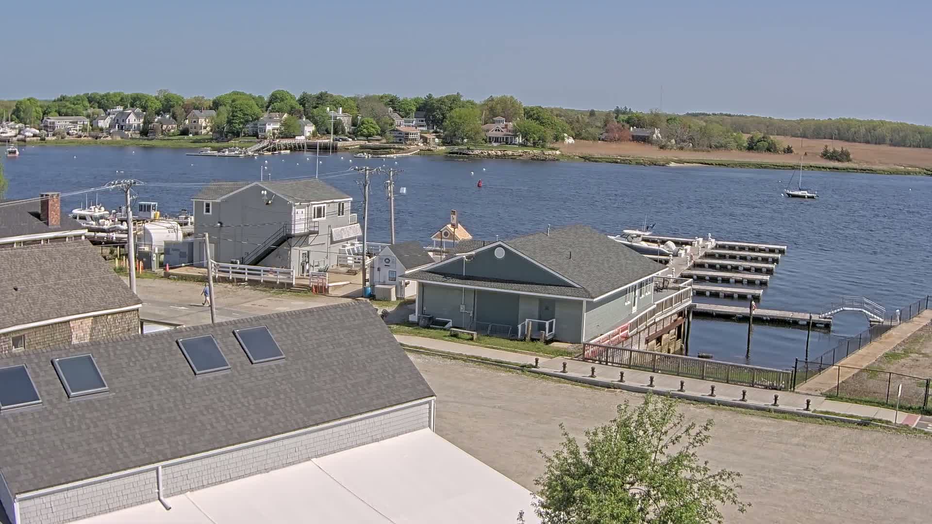 A waterfront scene on a sunny day shows a small town with houses, boats docked at a marina, and a calm river with a sailboat in the distance.
