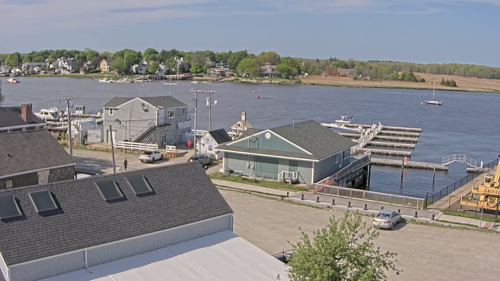 A waterfront view on a partly sunny day shows several buildings, boats docked at a marina, and cars parked in a lot, all situated next to a calm body of water.